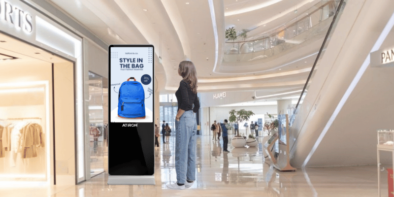 Digital display kiosk in a shopping mall showing a blue backpack advertisement with the text “Style in the Bag,” person viewing nearby.